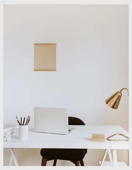 a light grey laptop on a white office desk, a golden coloured desk lamp and a black seat in front of a white wall for content creation services